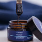 A close-up of a Trace Minerals Shilajit Resin jar showcases thick, dark resin rich in fulvic acid being lifted from the brown container. The blue label reflects its roots in Ayurvedic healing, with the lid set nearby.