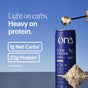A container of Ora Organic's So Lean & So Clean: Organic Vegan Protein Powder sits on a reflective surface with a scoop above it. Text shows: Light on carbs. Heavy on protein. 1g net carbs*, 23g protein* per serving.