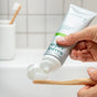 A hand squeezes ATTITUDE super leaves Fluoride-free Toothpaste onto a bamboo toothbrush, against a white tiled wall with a cup and another toothbrush in the background.