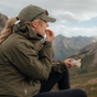 A person in an olive green jacket and cap sits outdoors in the mountains, enjoying a partially unwrapped Kates Real Food Organic Protein Bar - Snickerdoodle, fueling up with balanced energy while taking in the scenic view.