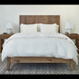 A neatly made bed featuring the Coyuchi Organic Crinkled Percale™ Duvet Cover and two pillows, flanked by wooden nightstands with matching lamps, sits atop a patterned rug in a simple, modern bedroom.