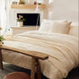 A cozy bedroom features a neatly made bed adorned with the Coyuchi Organic Crinkled Percale™ Duvet Cover, layered over cream linens and breathable bedding. A rustic wooden bench sits at the foot of the bed, surrounded by potted plants and softly glowing lights. Sheer curtains allow natural light to filter in through the window, while the organic cotton duvet cover from Coyuchi adds both comfort and style to this serene retreat.