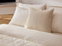 A neatly made bed with cream-colored sheets, a textured beige throw pillow, and two white pillows against a white headboard in a softly lit bedroom, featuring breathable bedding and enhanced comfort with the Coyuchi Organic Crinkled Percale™ Duvet Cover.