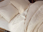 A neatly made bed with Coyuchi’s Organic Crinkled Percale™ Sheet Set in cream, complemented by matching organic cotton pillows and a comforter, is topped with a textured beige cushion. The breathable bedding looks soft and cozy.
