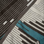 A close-up of the Coyuchi Morelia Organic Beach Towel, featuring a geometric pattern with black, gray, white, and teal stripes. A small fabric tag is partially visible in the top right corner.