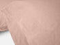 Close-up of a soft pink pillowcase with subtle stitching detail along the edge, lying on a bed with matching fabric. The texture is smooth and the bedding appears lightweight and breathable, crafted from GOTS certified cotton as part of Coyuchi’s Organic Crinkled Percale™ Sheet Set.