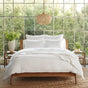 A modern bedroom features a wooden bed topped with Coyuchi’s Organic Crinkled Percale™ Duvet Cover, offering white, breathable comfort. Large windows reveal lush greenery outside, while potted plants and neutral furniture enhance the tranquil atmosphere. A woven light fixture hangs overhead, adding warmth to the serene space.