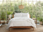 A cozy bedroom with a large window lets in natural light. The bed is adorned with crisp white Organic Crinkled Percale™ Pillowcases from Coyuchi, made with GOTS certified cotton. Green plants and wooden furniture surround the space, creating a fresh and inviting atmosphere.