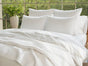 A neatly made bed with white bedding and Coyuchi’s Organic Crinkled Percale™ Pillowcases is shown near a large window overlooking greenery. A vase filled with green flowers rests on the nightstand.