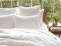 A neatly made bed with white bedding and Coyuchi’s Organic Crinkled Percale™ Pillowcases sits next to a wooden nightstand holding a green plant, a glass of water, and books. Large windows in the background reveal green trees outside.