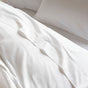 A close-up view of a neatly made bed with white sheets, pillowcases, and a textured blanket at the edge, featuring the Coyuchi Organic Crinkled Percale™ Duvet Cover for a breathable and effortlessly stylish finish.