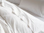 A close-up view of a neatly made bed with breathable bedding, featuring Coyuchi’s Organic Crinkled Percale™ Sheet Set in crisp white and pillowcases, completed by a textured cream-colored blanket at the edge.