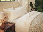 A neatly made bed with white pillows and an Organic Crinkled Percale™ Sham by Coyuchi, topped with a floral-patterned comforter. The breathable cotton sham adds a touch of comfort, while a wooden nightstand holds a lamp, flowers, and glasses. Sunlight streams through large windows with plants nearby.