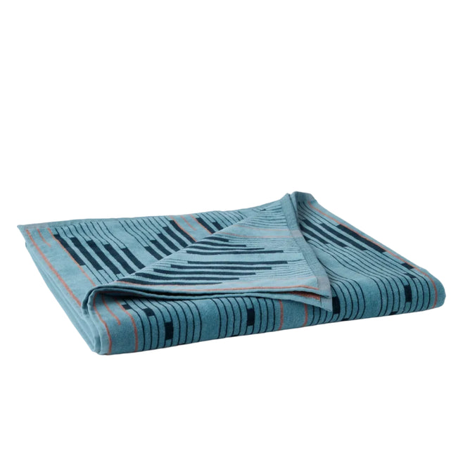 The Coyuchi Morelia Organic Beach Towel, shown folded against a plain white background, features an absorbent blue cotton fabric with a geometric pattern of dark and light lines accented by thin orange stripes.