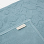 A close-up of the Wildflower Organic Bath Rug by Coyuchi, showcasing its textured, light blue surface adorned with a vintage-inspired floral pattern. A small fabric tag displays the Coyuchi brand name, emphasizing the rug’s absorbent quality and stylish design.
