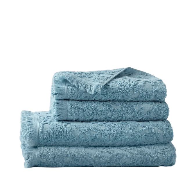 Four neatly folded Coyuchi Wildflower Organic Bath Towels, featuring textured mid-century floral patterns in blue, are stacked on a white background. Made from organic cotton, each towel showcases a subtle wildflower design. The top towel has one corner turned over to reveal the underside.