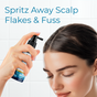 A woman with dark hair sprays Tree To Tub's Instant Relief Calming Scalp & Hair Spray for Dry, Itchy or Flaky Scalp onto her scalp against a white tiled background. Blue text above reads, Spritz Away Scalp Flakes & Fuss.