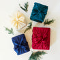 Four boxes wrapped in Sarahs Silks Holiday Reusable Gift Wrap Furoshiki Set (cream, teal, navy, red) are arranged on a white surface with pine branches, highlighting sustainable and beautiful gift wrapping.