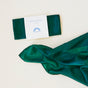A folded green silk cloth from Sarahs Silks, labeled Holiday Reusable Gift Wrap Furoshiki Set, sits beside an unfurled section of the same fabric—ideal as sustainable gift wrap or elegant Furoshiki—on a light background.