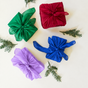 Four Holiday Reusable Gift Wrap Furoshiki Set gift boxes wrapped in colorful silk with knotted tops, arranged with sprigs of evergreen on a white background.