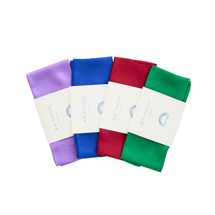 Holiday Reusable Gift Wrap Furoshiki Set containing four folded Mini Playsilk squares in lavender, blue, red, and green, each secured with a paper band.