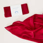 A folded red Holiday Reusable Gift Wrap Furoshiki Set wrapped with a paper band labeled Mini Playsilk Seasonal next to an unfolded red play silk.