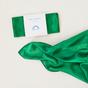 Two folded pieces of green silk fabric for a Holiday Reusable Gift Wrap Furoshiki Set, one contained in a white paper wrapper labeled Mini Playsilk Seasonal with a rainbow icon.