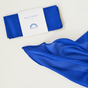 A folded blue silk Holiday Reusable Gift Wrap Furoshiki in its packaging next to an identical unfolded blue silk square.