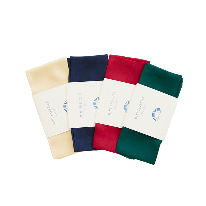 The Holiday Reusable Gift Wrap Furoshiki Set by Sarahs Silks features four folded bandanas in cream, navy blue, red, and green with “Mini Parisik” rainbow paper bands, perfect as sustainable gift wrapping, shown on a white background.