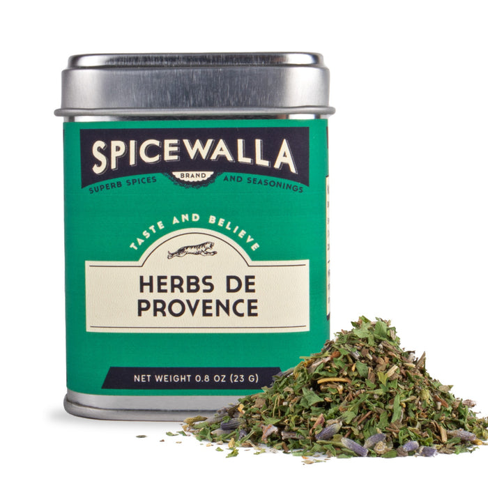 A green-labeled tin of Spicewalla Herbs de Provence (0.8 oz/23 g) sits beside a small pile of dried mixed herbs, its silver lid and product label displaying the brand and contents.