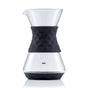 The Saki Pour-Over Glass Coffee Maker features a clear borosilicate carafe with a geometric black silicone grip and is ideal for pour-over coffee. Its wide bottom and narrow neck ensure optimal brewing, and “SAKI” is labeled on the front.