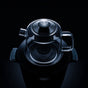 A glass teapot with lid and handle rests atop the SAKI Electric Samovar, both adorned with sleek stainless steel accents, set against a dark, dramatic background.