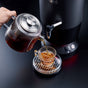 A hand pours tea from a glass teapot into a clear cup on a metal tray, next to the sleek SAKI Electric Samovar with a spout, ensuring every pour is smooth and bitterness-free.