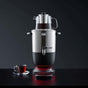 The SAKI Electric Samovar, featuring a stainless steel tea maker and a glass teapot on top filled with smooth, bitterness-free tea, stands elegantly. Beside it on a black surface and against a dark background sits a glass cup and saucer filled with freshly brewed tea.