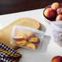 Sliced peaches are stored in a Stasher Reusable Silicone Sandwich bag (2-pack) on a white countertop, surrounded by whole peaches in a bowl, granola in another bag, a wooden cutting board, and a checkered cloth.