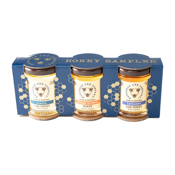 Three jars of Orange Blossom Honey, Acacia Honey, and Lavender Honey are presented in the Savannah Bee Company Honey Artisanal Sampler Set—a blue box with honeycomb designs. Each jar features a white label and a gold lid.