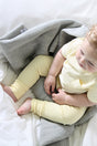 A baby in a pale yellow shirt and ribbed leggings sits on a personalized The Sawa - Moon Stone baby blanket, partially covered by a gray textured throw.