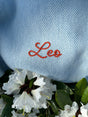 The Sawa - Sky Blue merino baby blanket embroidered with the name Leo in orange cursive thread, resting above white flowers.