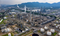 Aerial view of an industrial oil refinery with towers, tanks, and pipelines emitting smoke—underscoring plastic pollution concerns—promotes Grove Co.'s The Unplastic Your Life Educational Campaign against a backdrop of green hills and a hazy sky.