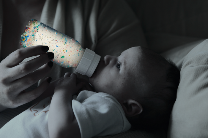As part of Grove Co.'s Unplastic Your Life Educational Campaign, a baby is bottle-fed under moody lighting, gazing up at a bottle filled with colorful fragments resembling plastic—symbolizing the dangers of plastic pollution.