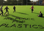 Four children play soccer on a green field reading “NO PLACE FOR PLASTICS,” promoting Grove Co.’s Unplastic Your Life Educational Campaign against plastic pollution. Goalposts and trees are visible under a partly cloudy sky.