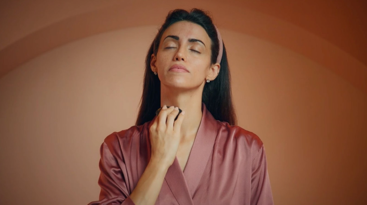 A woman in a rose-colored satin robe gently rolls a facial massage tool on her neck, with her eyes closed and a calm expression, against a warm orange background.