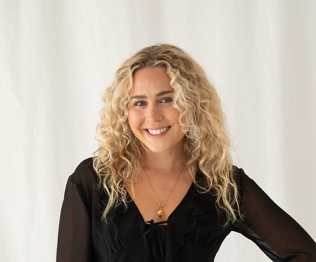 A woman with long, curly blonde hair smiles at the camera. She is wearing a black, sheer-sleeved blouse and a gold necklace. The background is a plain white curtain.