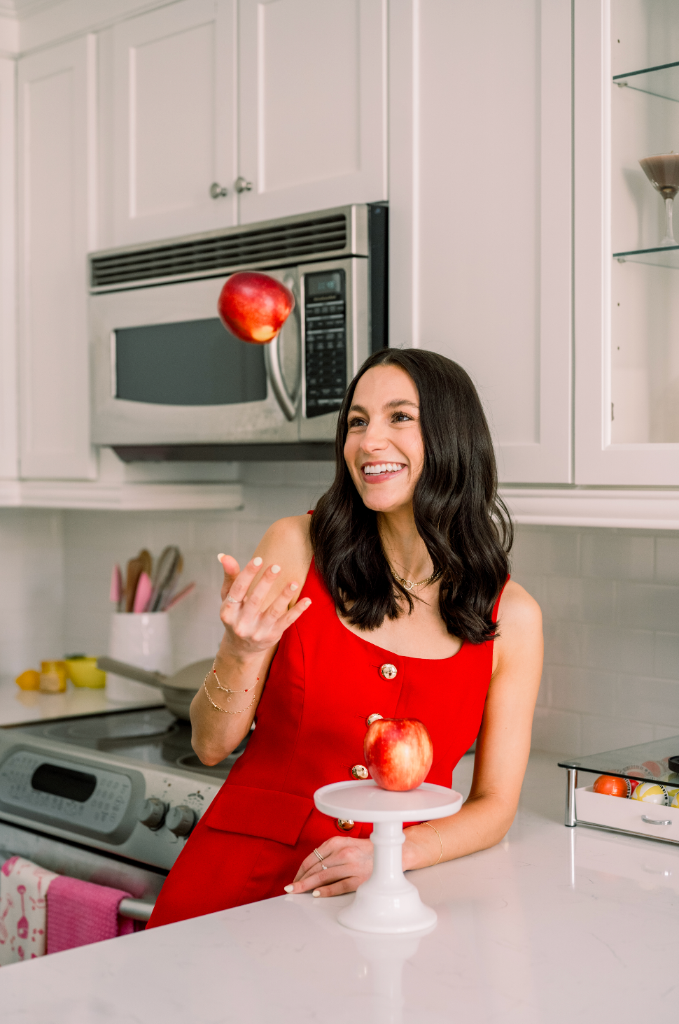 A woman in a bright red dress smiles in a kitchen while tossing an apple in the air. Another apple rests on a white cake stand in front of her. The kitchen has white cabinets and modern appliances.