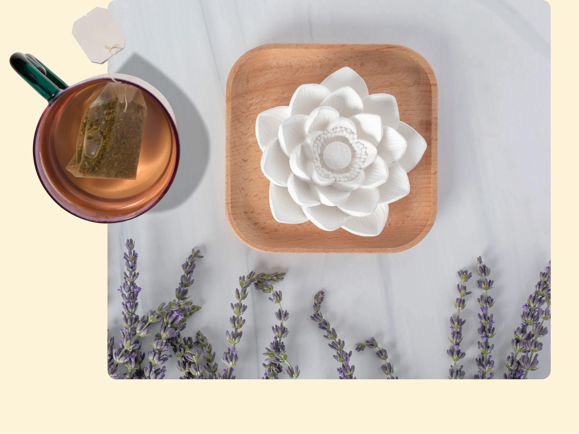 A cup of tea with a teabag sits beside a wooden tray holding a white flower-shaped object, with sprigs of lavender scattered on a marble surface.