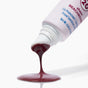 A tube of The Seaweed Bath Co Tinted Mineral Lip Shine SPF 20 in sheer beachberry, infused with nourishing shea butter, with a dark berry-colored product pouring out onto a white surface.