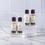 Tree To Tub’s Relaxing Lavender Travel Essentials Set features four mini bottles, including sulfate-free, vegan shampoo, displayed on clear stands against a white tiled background.