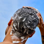 A person washes their short hair with Phil's Strengthening + Thickening Shampoo, lathering up white bubbles on their scalp with both hands, aiming for thicker hair against a blue sky backdrop.