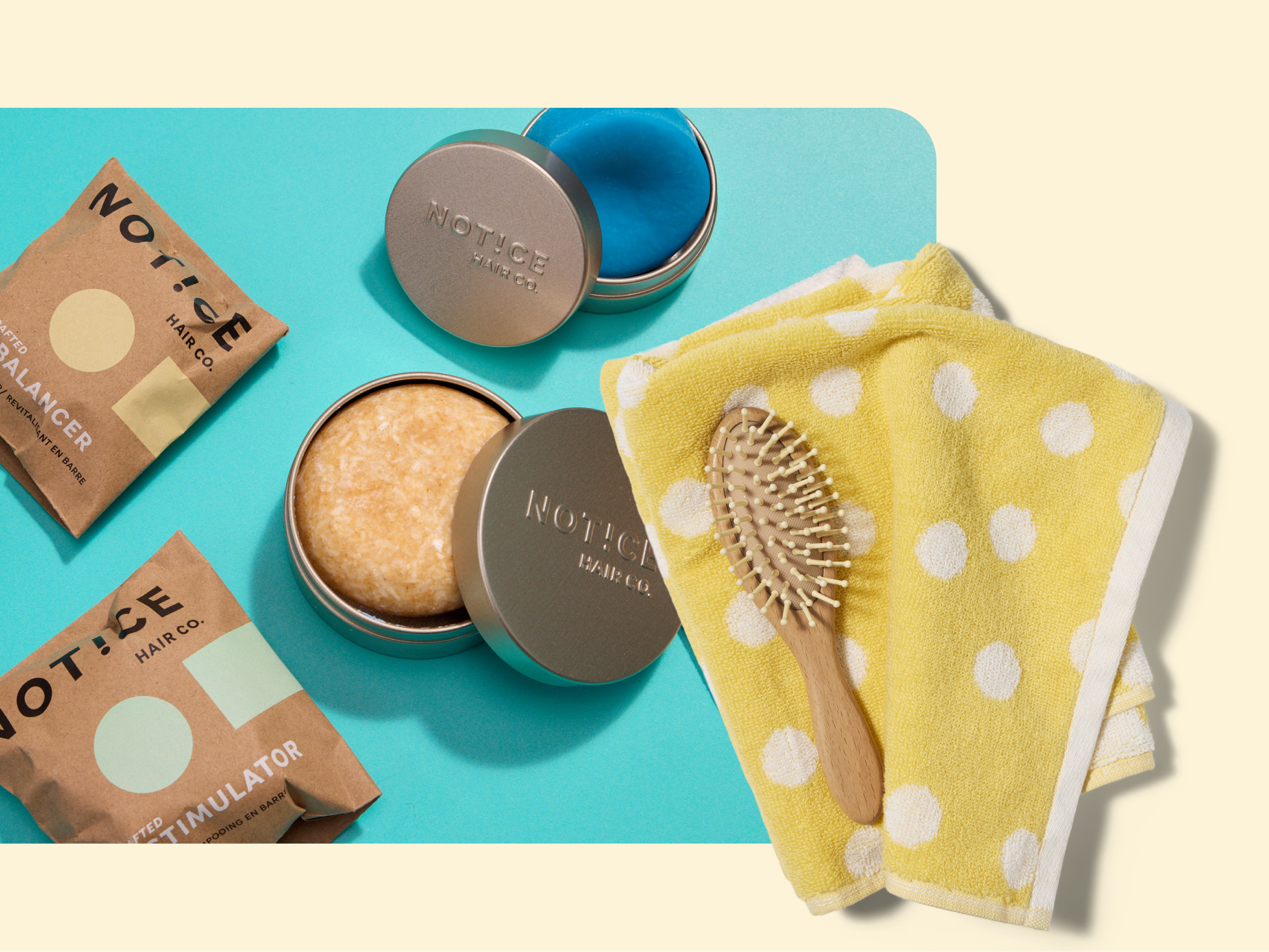 A yellow polka dot towel with a wooden hairbrush beside two tin containers with solid hair products and two cardboard hair product packets on a blue and cream background.