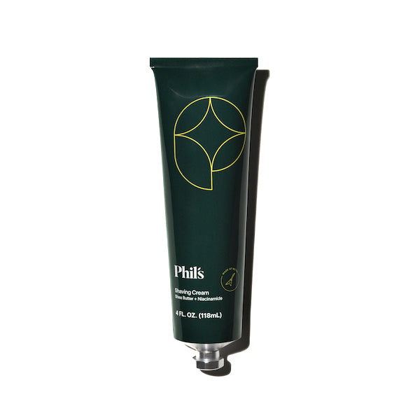 A dark green tube of Phils Shaving Cream with minimalist yellow line art and white text, featuring soothing shea butter, is shown slightly squeezed on a white background with a shadow to the right.